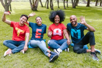Four people sitting outside on grass. They are wearing lettered t-shirt that spells team.